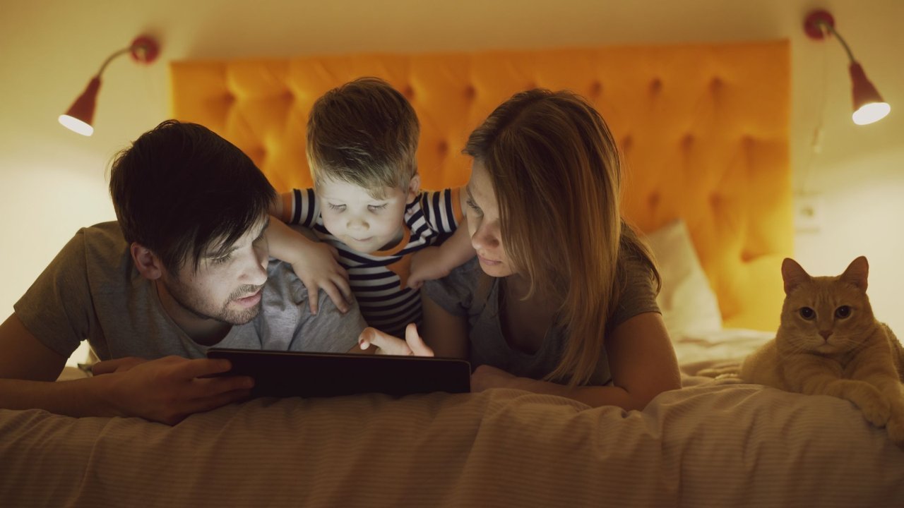 Pai, mãe e filho olham juntos a tela de um tablet na cama, com um gato ao lado